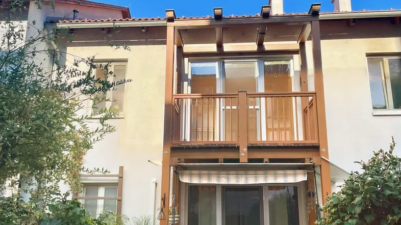 Außenansicht des Gebäudes mit einem Holzbalkon im Obergeschoss und einer überdachten Terrasse im Erdgeschoss.
