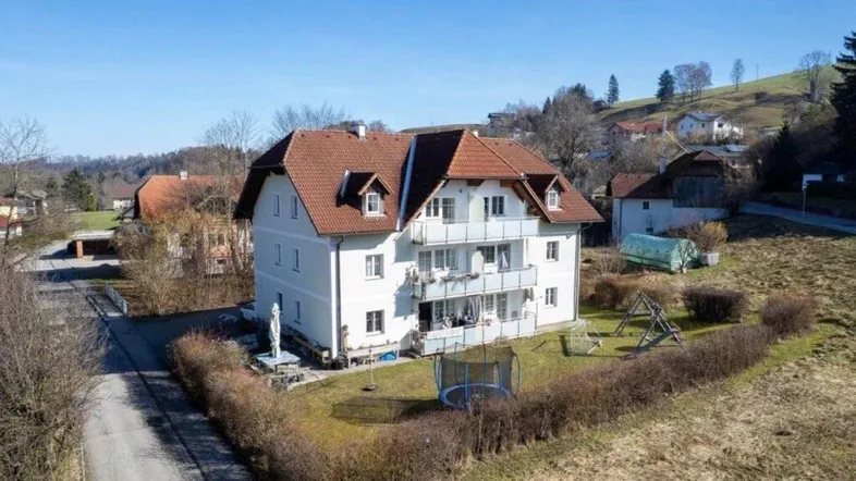 Mehrfamilienhaus mit Satteldach, Balkonen und großem Garten mit Spielgeräten in ländlicher Umgebung.