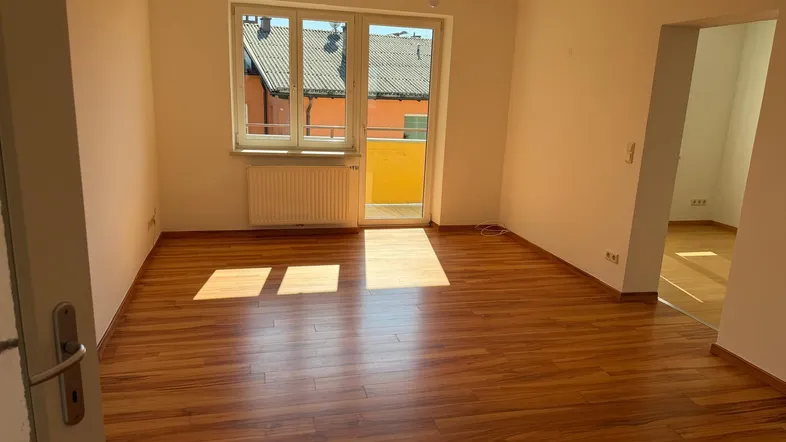 Helles, leeres Zimmer mit Holzboden und Zugang zu einem kleinen Balkon, ideal als Wohnbereich.