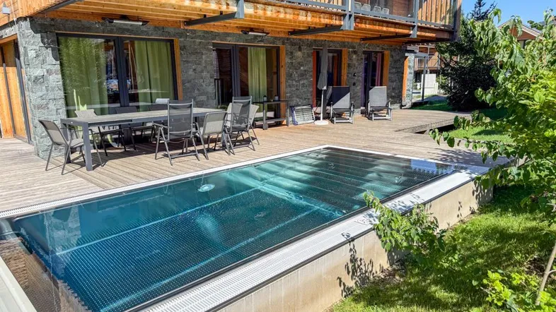 Große Holzterrasse mit Esstisch, Liegestühlen und einem eingelassenen Pool vor der modernen Fassade.