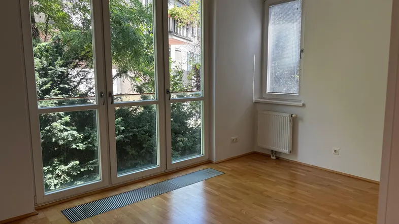 Lichtdurchfluteter Raum mit Holzparkett und großen Fenstern mit Blick auf üppiges Grün.