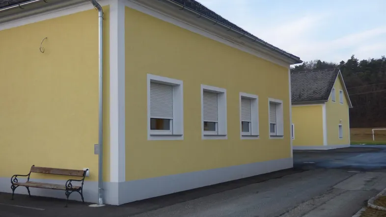 Gelbes Gebäude mit weißen Fenstern und Rollläden, umgeben von einer asphaltierten Fläche und einem Holzbänkchen.