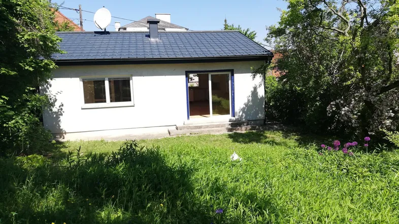Kleines Haus mit weißer Fassade, dunklem Dach und großem, grünem Gartenbereich.
