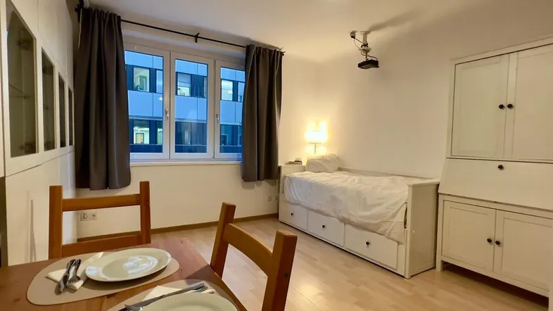 Helles Zimmer mit Esstisch, Schlafbereich und Blick auf umliegende Gebäude durch große Fenster.
