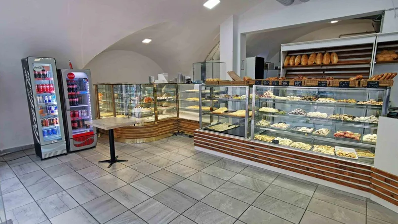 Geräumiger Verkaufsraum einer Bäckerei mit zahlreichen Auslagen, Kühlregalen und gefliestem Boden unter einem Gewölbedach.