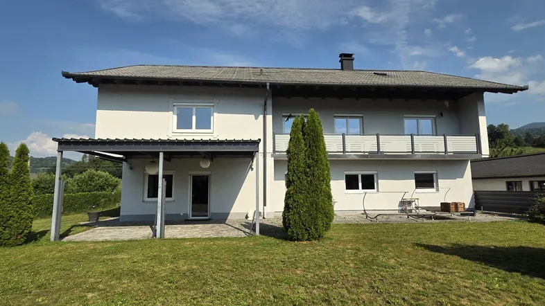 Hausrückseite mit großem Rasenbereich, überdachter Terrasse und Blick in die grüne Landschaft.