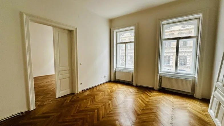 Helles Zimmer mit Fischgrätparkett und hohen Fenstern, das zu einem weiteren Raum führt.