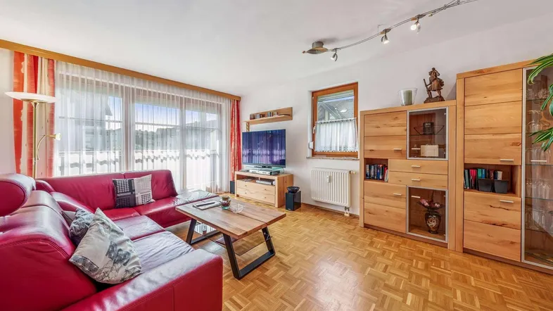 Helles Wohnzimmer mit rotem Sofa, Holzmöbeln und Parkettboden sowie Zugang zum Balkon.