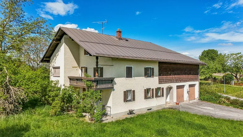 Traditionelles Landhaus mit Satteldach und Balkon, umgeben von grüner Landschaft unter blauem Himmel.