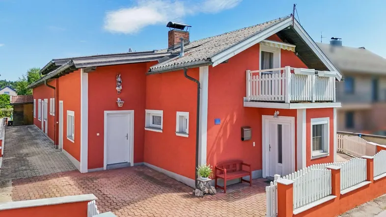 Rotes Einfamilienhaus mit weißem Zaun, kleinem Balkon und Satteldach unter blauem Himmel.