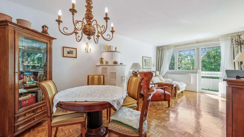 Helles Wohn- und Esszimmer mit Parkettboden, Kachelofen und Zugang zum Balkon.