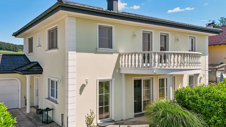 Einfamilienhaus mit heller Fassade, Balkon und gepflegtem Vorgarten unter blauem Himmel.