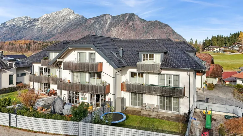 Modernes Mehrfamilienhaus mit Balkonen, Garten und Spielplatz vor beeindruckender Bergkulisse.