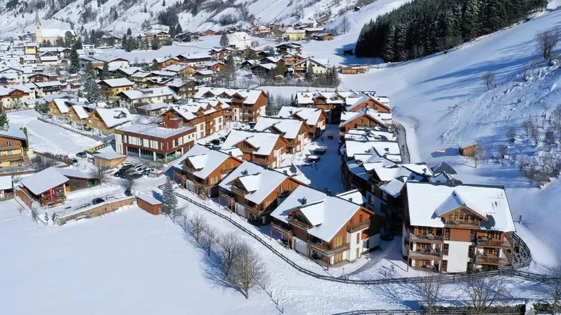 Luftaufnahme eines schneebedeckten Dorfes mit zahlreichen traditionellen Chalets und verschneiten Bergen im Hintergrund.