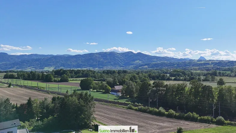 Weitläufige Landschaftsaufnahme mit Bergen im Hintergrund und Feldern im Vordergrund unter blauem Himmel.
