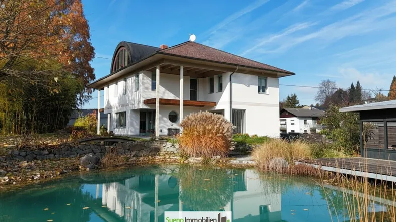 Großzügiges Einfamilienhaus mit weitläufigem Garten und idyllischem Schwimmteich unter blauem Himmel.