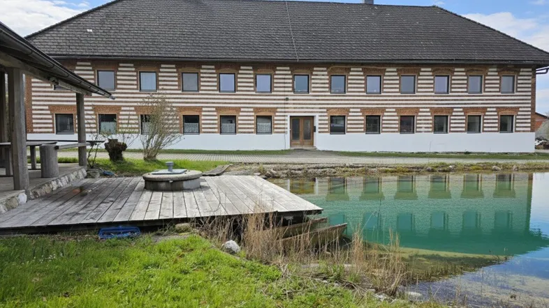 Großes Bauernhaus mit traditioneller Fassade, umgeben von einem Teich mit Holzsteg und ländlicher Umgebung.