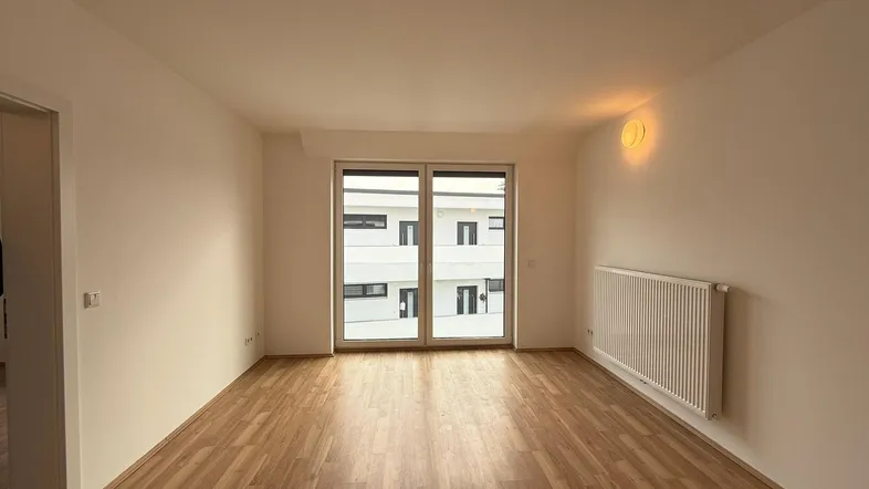 Helles, leeres Wohnzimmer mit Laminatboden und Zugang zu einem Balkon oder einer Terrasse.