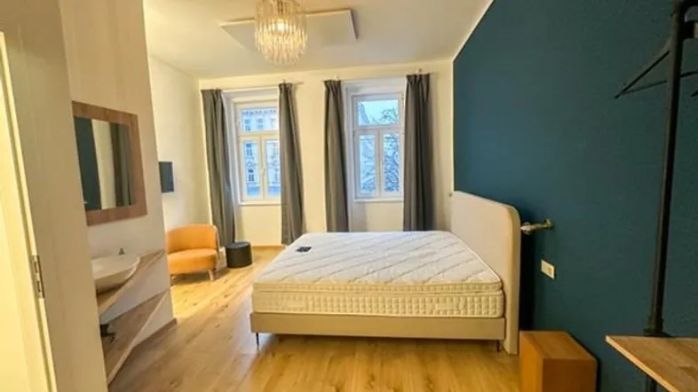 Schlafzimmer mit Doppelbett, Holzboden und einer dunkelblauen Akzentwand, inklusive kleinem Waschbereich.