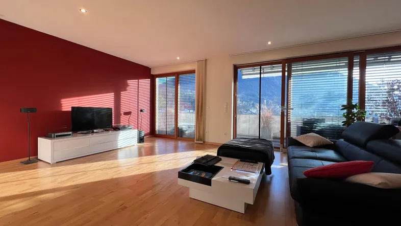 Helles Wohnzimmer mit Holzboden, roter Akzentwand und Zugang zum Balkon mit Bergblick.