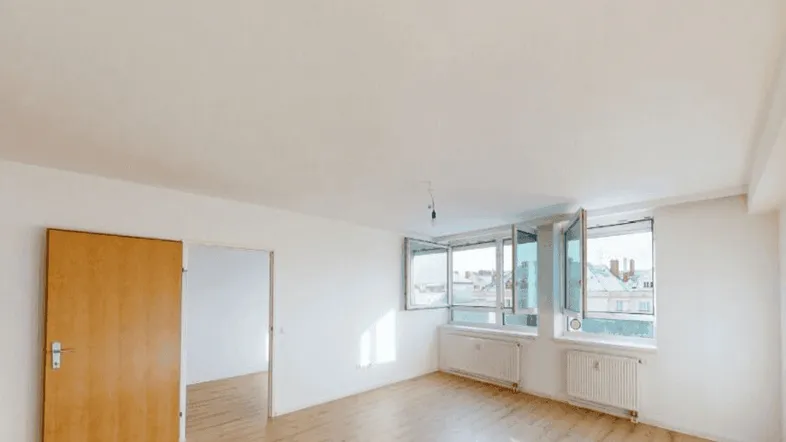 Helles, leeres Wohnzimmer mit Parkettboden und großen Fenstern, die viel Tageslicht hereinlassen.