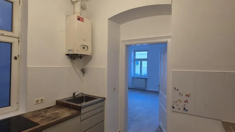 Einfache Küche mit Spüle, Kochfeld und Gastherme, die zu einem weiteren Raum führt.