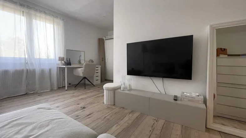 Helles Wohnzimmer mit Holzboden, Fernseher und einem Arbeitsbereich am Fenster.