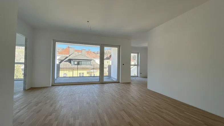 Heller, leerer Wohnbereich mit neuem Parkettboden, großen Fenstern und Zugang zu einem Balkon mit Stadtblick.