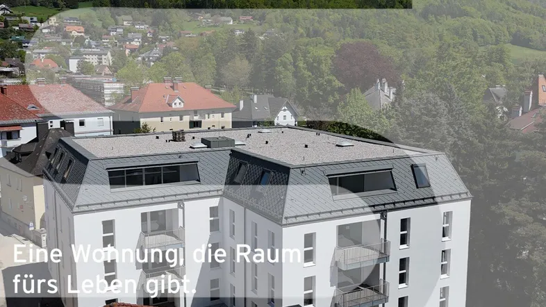 Modernes Mehrfamilienhaus mit Balkonen und Mansarddach in grüner Hügellandschaft mit Bergblick.