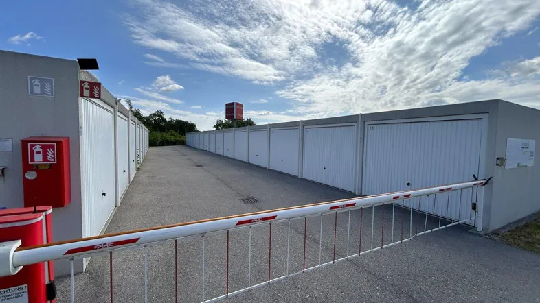 Einfahrt zu einer Reihe von Garagenboxen mit Schranke und Sicherheitsmerkmalen unter blauem Himmel.