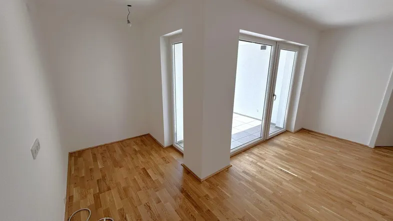 Helles Zimmer mit Parkettboden und Zugang zu einem Außenbereich durch große Glastüren.