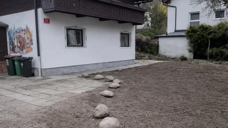 Hausfassade mit traditionellem Holzbalkon und einem vorbereiteten Gartenbereich unter bewölktem Himmel.