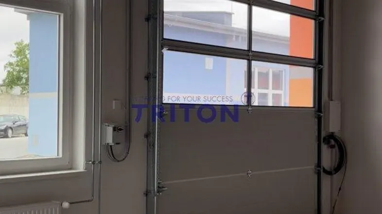 Innenansicht einer Gewerbehalle mit großem Fenster, Rolltor, Betonboden und sichtbaren Holzträgern.