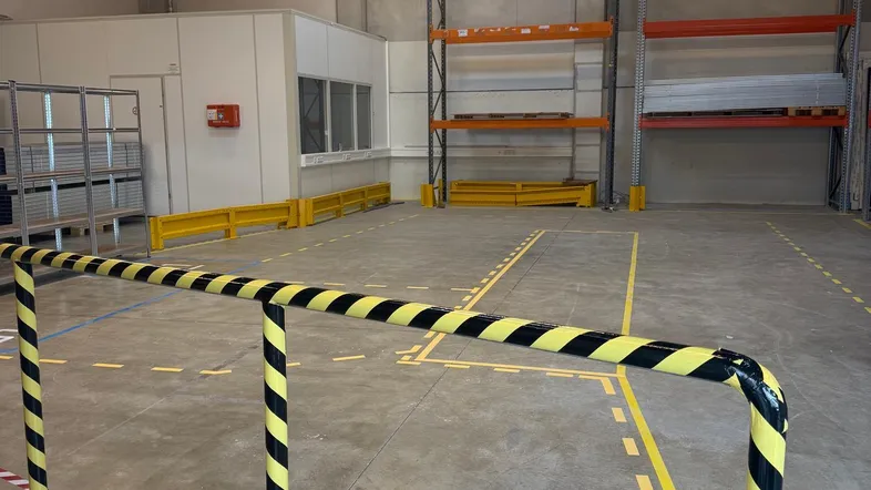 Geräumige Lagerhalle mit Betonboden, hohen Decken und gelb-schwarzen Sicherheitsbarrieren für den Betrieb.
