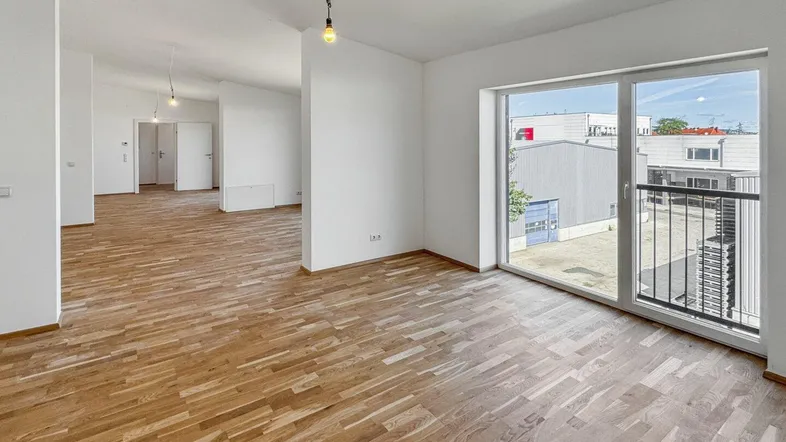Geräumiger, leerer Wohnbereich mit hellem Holzboden und großen Fenstern, die viel Tageslicht hereinlassen.