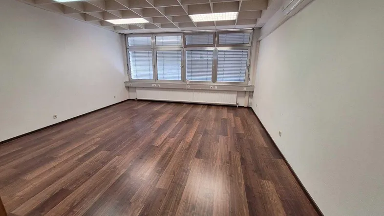 Leerer Büroraum mit dunklem Holzboden, weißen Wänden und großen Fenstern mit Jalousien.
