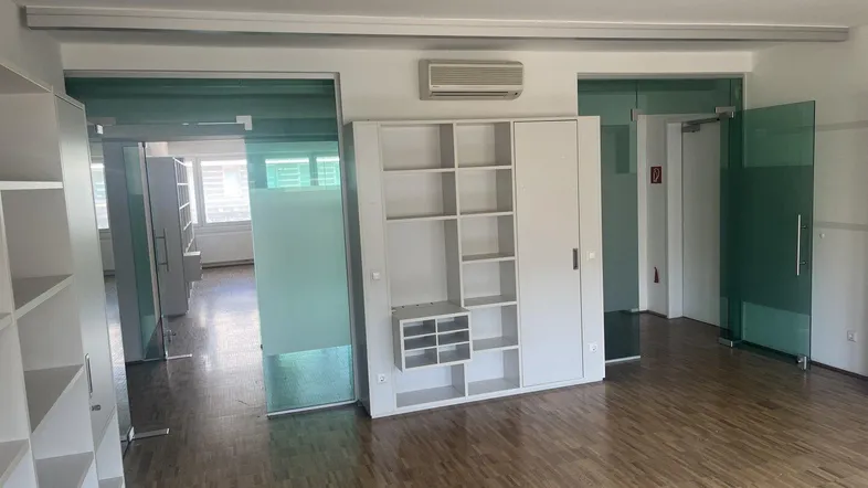 Geräumiges Büro mit Holzparkettboden, weißen Regalen und modernen Glastüren, die zu weiteren Räumen führen.