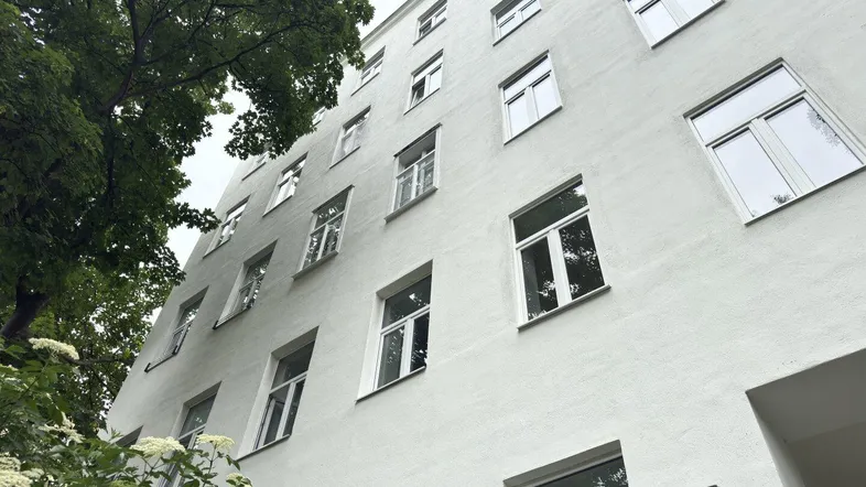 Weiße Gebäudefassade eines Mehrfamilienhauses mit vielen Fenstern und umgebenden Bäumen.