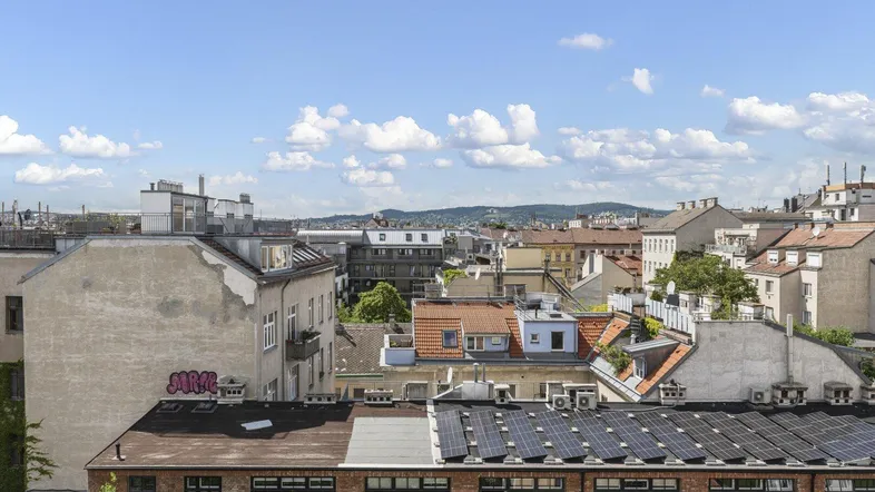 Panoramablick über die städtischen Dächer mit Solaranlagen und grünen Hügeln am Horizont unter blauem Himmel.