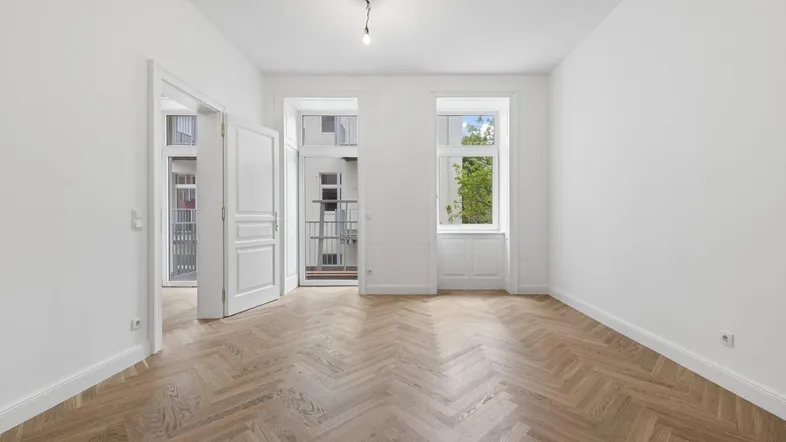 Heller Raum mit Fischgrätparkett, weißen Wänden und Zugang zu einem französischen Balkon.