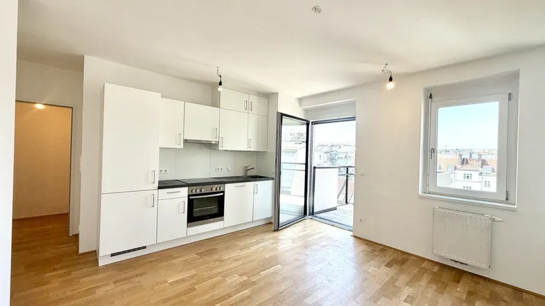 Heller offener Wohnbereich mit integrierter Küche, Holzboden und Zugang zum Balkon.