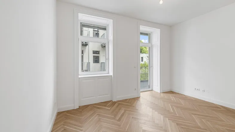 Heller Raum mit elegantem Fischgrätparkett, hohen Decken und Zugang zu einem Balkon.