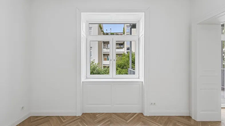 Heller Raum mit Fischgrätparkett, weiß gestrichenen Wänden und Blick ins Grüne durch das Fenster.