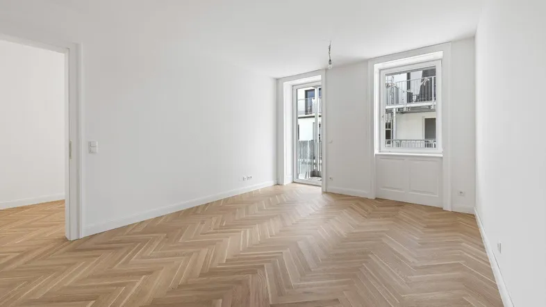 Heller Raum mit Fischgrätparkett, weißen Wänden und Zugang zu einem französischen Balkon.