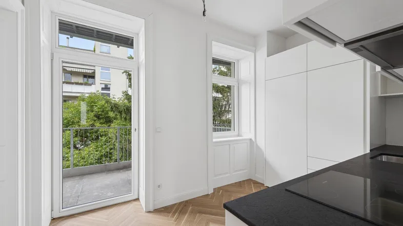 Moderne Küche mit weißen Schränken, dunkler Arbeitsplatte und Fischgrätparkett, mit Zugang zum Balkon und Grünblick.