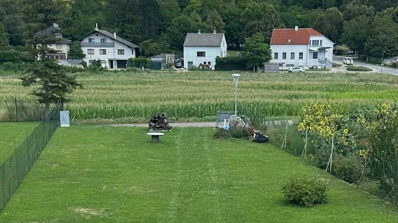 Blick über einen gepflegten Rasen auf ein Feld und Wohnhäuser in ländlicher Umgebung unter blauem Himmel.