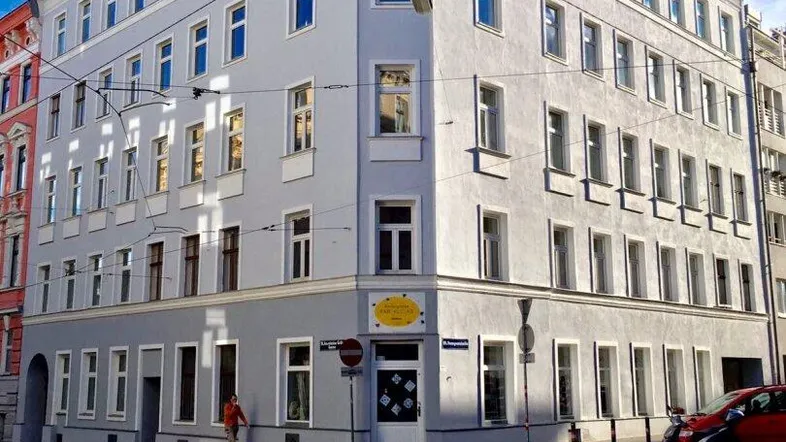 Eckgebäude in städtischer Umgebung mit klassischer Architektur und vielen Fenstern unter blauem Himmel.