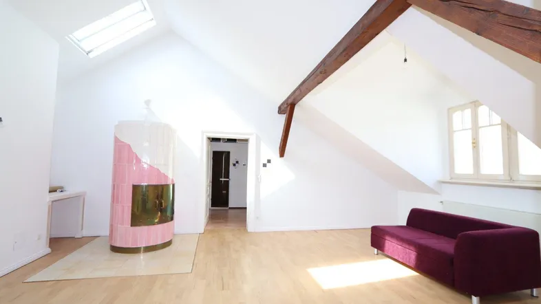Helles Dachgeschosszimmer mit sichtbaren Holzbalken, Kachelofen und Dachfenster, ausgestattet mit einem Sofa.