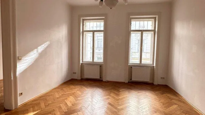Helles Zimmer mit Fischgrätparkett, hohen Fenstern und einem klassischen Kronleuchter.