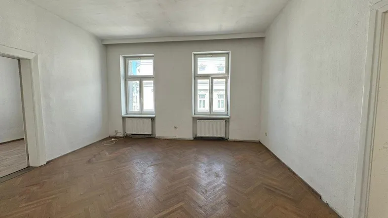 Helles Zimmer mit Fischgrätparkett und zwei Fenstern, bereit zur Renovierung.